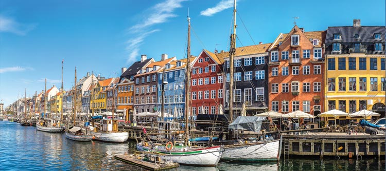 Colorful houses line the water in Copenhagen, Denmark. Several white sailboats are docked.